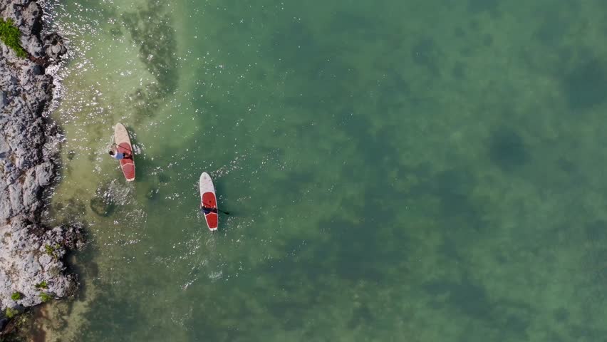 Two people riding paddle boards on vacation and Crystal clear blue water near racks on an island in Mexico overhead aerial drone epic cinematic