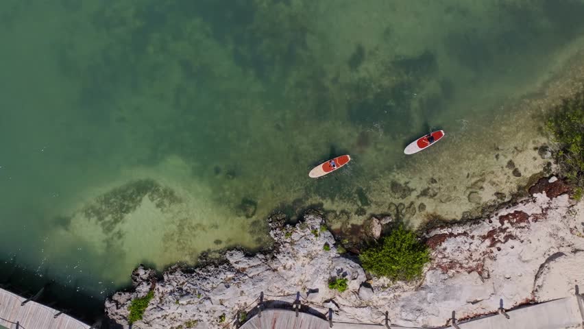 Two people riding paddle boards on vacation and Crystal clear blue water near racks on an island in Mexico overhead aerial drone epic cinematic