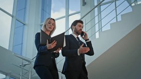 Two business professionals walking down modern office staircase. Bearded executive talking smartphone giving documents to woman coworker. Busy partners going downstairs corporate building with folder - Powered by Shutterstock - Get 15% off with code: PIKWIZARD15