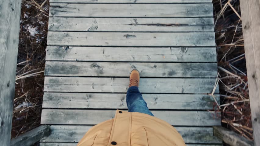 A POV Perspective Of A Man In Hiking Boots Walking Across A Wooden Suspension Bridge Over A Wild Mountain Lake. The View Highlights The Adventure And Thrill Of Outdoor Exploration.