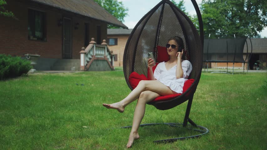 Happy, smiling young Caucasian woman barefoot in summer dress, sits on swing near village house, listens to music. Portrait of attractive woman in wireless headphones, sunglasses with phone in hands.