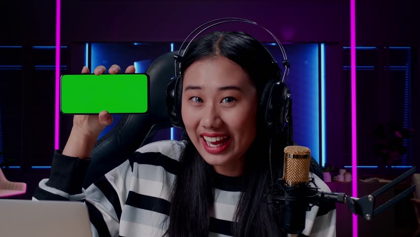 Close Up Of A Smiling Asian Woman With Headphone And Computer Reviewing Green Screen Mobile Phone In Room Lit By Neon Lights In Retro