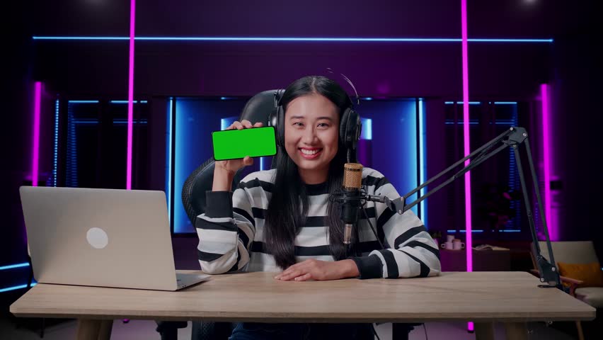 A Smiling Asian Woman With Headphone And Computer Showing Green Screen Mobile Phone In Room Lit By Neon Lights In Retro