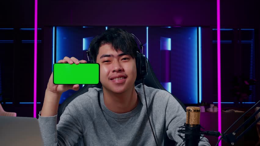 Close Up Of A Smiling Asian Man With Headphone And Computer Showing Green Screen Mobile Phone In Room Lit By Neon Lights In Retro