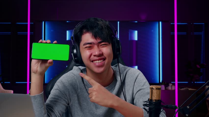 Close Up Of A Smiling Asian Man With Headphone And Computer Reviewing Green Screen Mobile Phone In Room Lit By Neon Lights In Retro