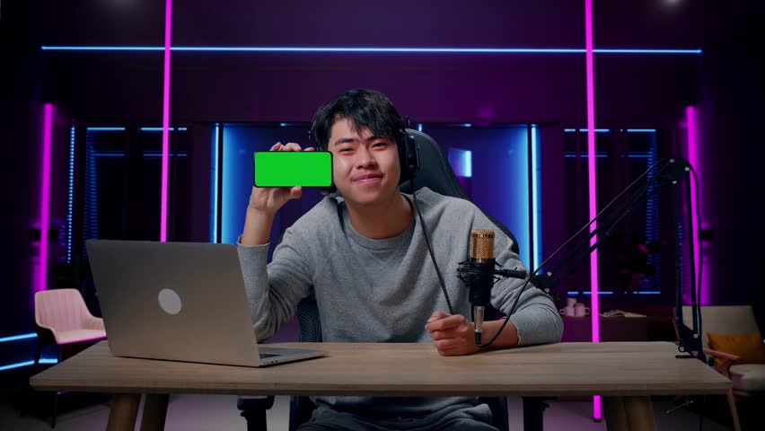 A Smiling Asian Man With Headphone And Computer Showing Green Screen Mobile Phone In Room Lit By Neon Lights In Retro