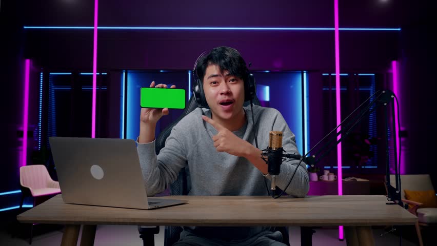 A Smiling Asian Man With Headphone And Computer Showing Green Screen Mobile Phone In Room Lit By Neon Lights In Retro