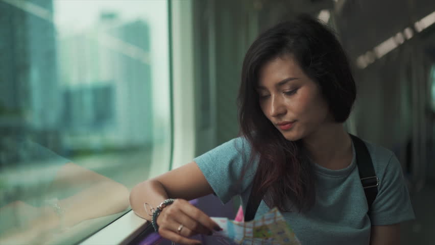 Cinematic Close Up shot of Young Woman Looking Through Window While Traveling by Train Through City Landscape. Map in Hands. Nostalgic Female Feeling Homesick and Smiling While Reaching her Station