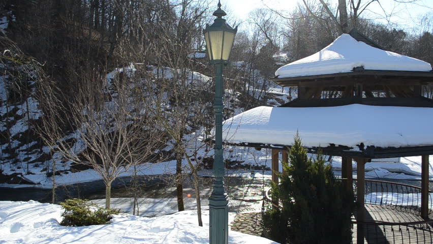 wooden bower roof covered with snow and retro lighting lamp pole on river water bank waterside in winter park.
