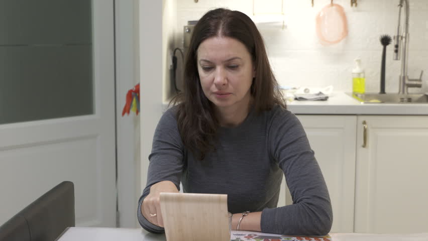 Adult woman with a swollen, tear-stained face at the kitchen table in the morning. The face of a sad, tired or sick woman reading messages on a smartphone screen.