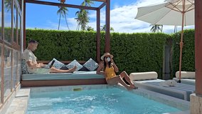 A successful freelancer works on his laptop beside a stunning pool villa in Thailand with palm trees swaying in the background. A diverse couple of man and woman at a pool villa - Powered by Shutterstock - Get 15% off with code: PIKWIZARD15