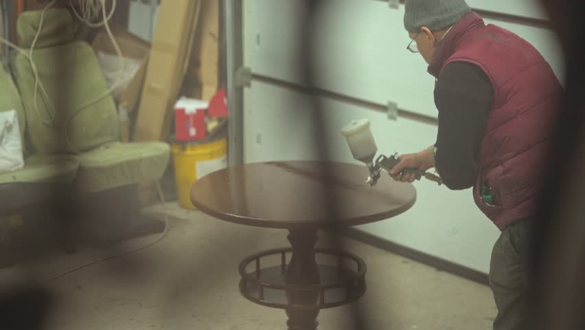 Hardworking senior male carpenter spraying varnish or primer using compressed air on a wooden tabletop a vintage table. Furniture restoration workshop Manual work Small business Carpentry Man at work.