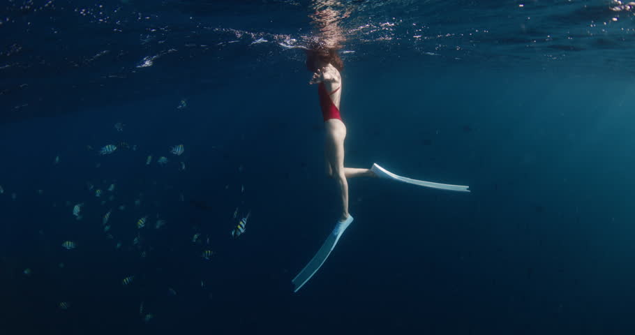 Woman swims in blue ocean with school of tropical fish, underwater view