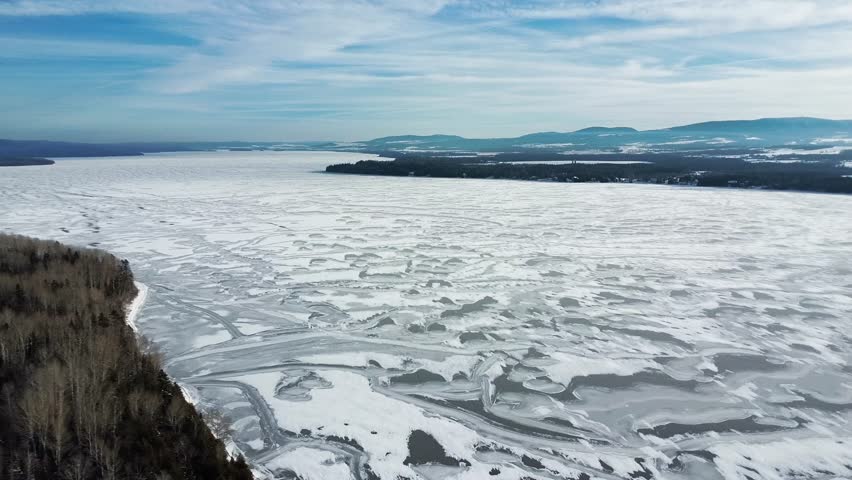 Panoramic aerial view of patterns on the ice of a vast frozen lake bordered by dense forests under a clear sky, an overview of a winter landscape. Matapedia Lake, quebec, Canada, 2024.