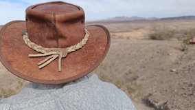 A person stands in a desert area, wearing a cowboy hat and a hoodie. They look at a warning sign about unstable rocks, surrounded by dry terrain and distant mountains. - Powered by Shutterstock - Get 15% off with code: PIKWIZARD15