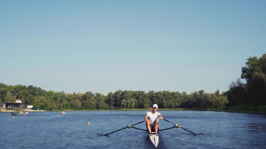 Rower solo training. Side view of young caucasian male rower, during a rowing practice, athlete sitting in a boat in the river Dnipro, rows through a calm water in autumn. 4k footage. Caucasian man tr