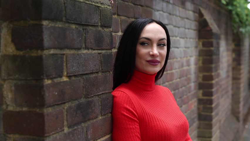 Brunette woman in a red turtleneck sweater leaning against a brick wall on a london street, smiling confidently while exuding an air of casual elegance and modern style