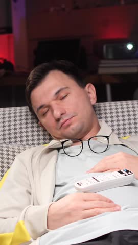 vertical video Man sleeping in armchair with remote control and glasses after watching tv