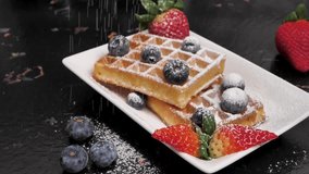Delicious waffles in a white plate on a black table with the addition of fresh strawberries and blueberries sprinkled with powdered sugar in slow motion. Belgian waffles for breakfast. - Powered by Shutterstock - Get 15% off with code: PIKWIZARD15
