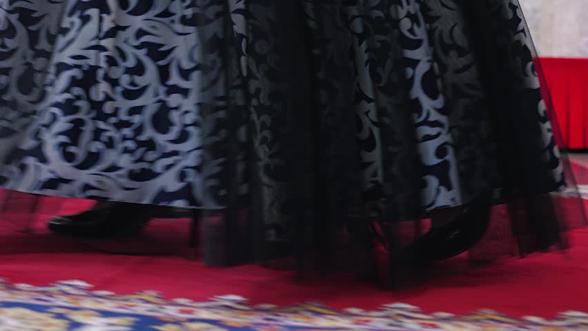 Close-up of a woman in a flowing black and white dress with intricate patterns walking down the red carpet. Black high-heeled shoes are visible under the dress. Side view. Slow motion