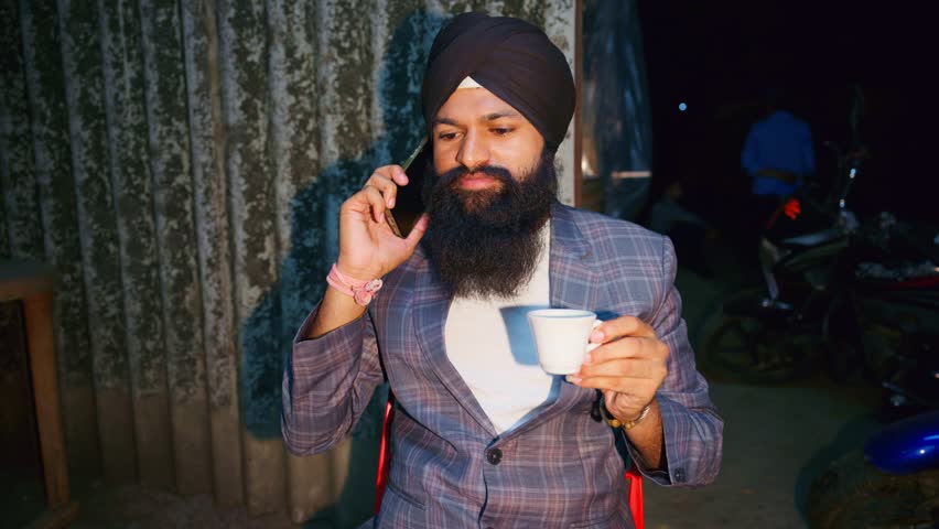 Turban person drinks coffee as speak over call, Punjabi man calling on phone while having tea, Bearded male engaged in conversation, He talk from mobile, Gentleman seated at a cafe table, Roadside