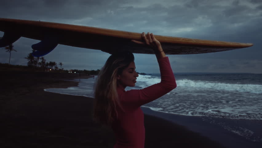 Young woman surfer walks along sand ocean beach, holds surfboard above head after sup training. Attractive girl walking or run to surfing with board. Evening dusk on bali, indonesia. Pink swuimsuit