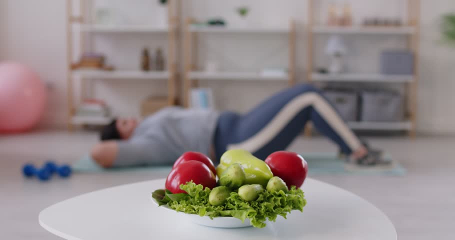 Close up of plate with healthy fresh vegetables with overweight woman doing abdominal crunches on blurred background. Comprehensive approach to weight loss, physical activity and healthy nutrition.