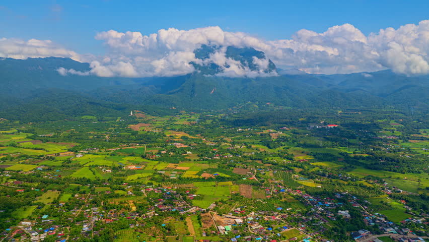 Arial hyper lapse view of Villages and rice fields surround the towering Doi Luang Chiang Dao. White clouds on the mountaintop contrast with the blue sky covering the changing shapes of the clouds.
