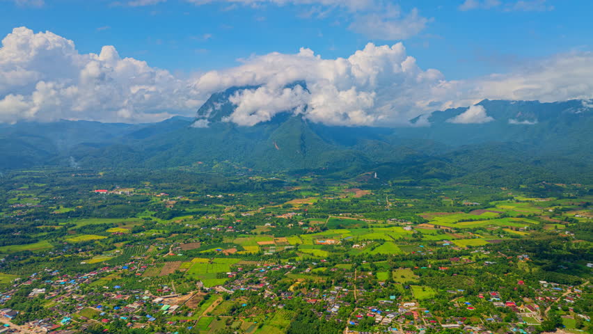 Arial hyper lapse view of Villages and rice fields surround the towering Doi Luang Chiang Dao. White clouds on the mountaintop contrast with the blue sky covering the changing shapes of the clouds.