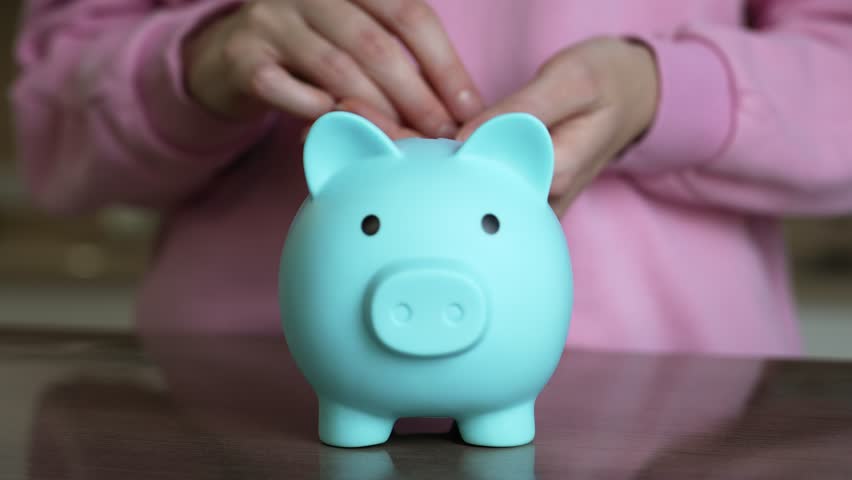 Young woman wearing a pink sweater places coins into a blue piggy bank. A dark table serves as the backdrop for this indoor scene focused on saving and financial planning