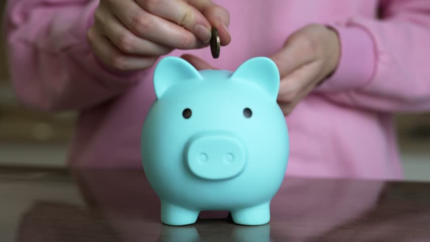 Young woman wearing a pink sweater places coins into a blue piggy bank. A dark table serves as the backdrop for this indoor scene focused on saving and financial planning