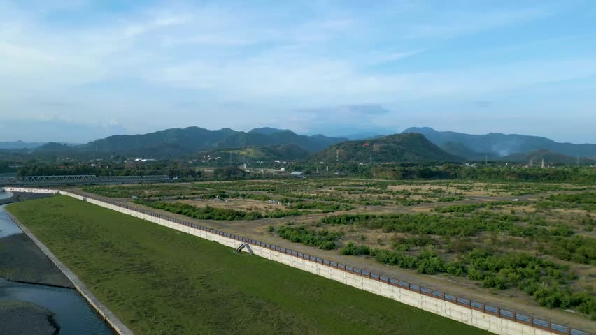 Aerial view of a vast landscape featuring rolling hills and a long concrete barrier. The scene captures the lush greenery and distant mountains under a clear blue sky, showcasing natural beauty