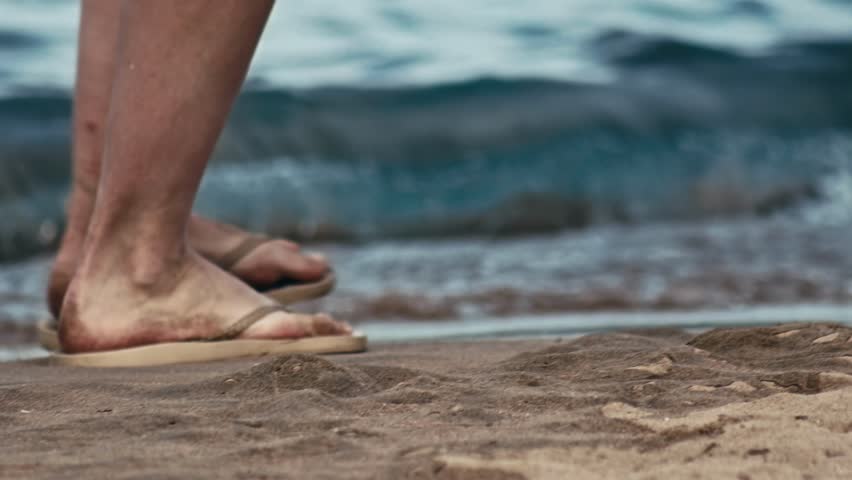 Male legs with flip-flops slippers walk along sandy beach by sea, close up shot of feet with sandals shoes. Stunning waterfront. Calm coastal waters are perfect for a cruise. Sharm El Sheikh, Egypt
