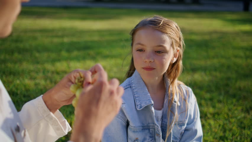 Small kid eating grape looking on unknown mother in summer picnic closeup. Unrecognizable woman feeding cute girl with tasty food. Portrait of blond child tasting delicious dessert on sunny weekend
