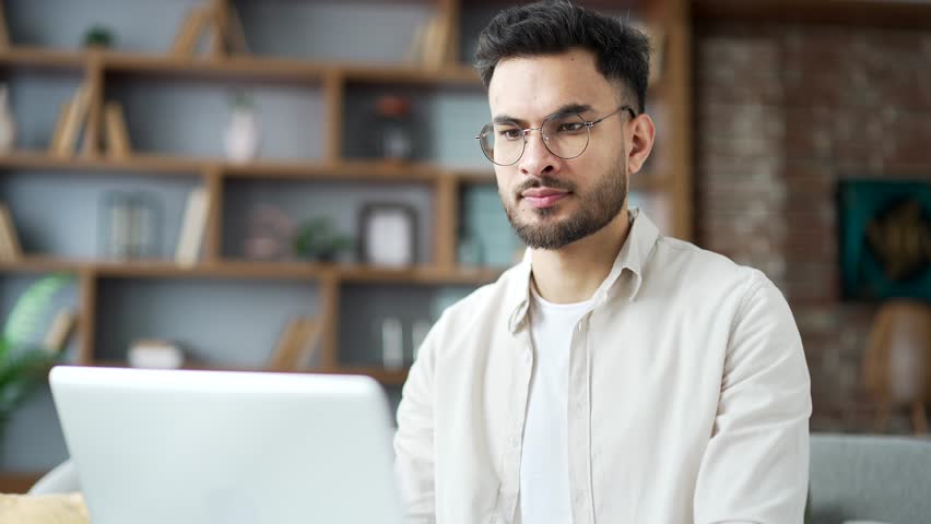 Young adult man wearing glasses working on laptop sitting on sofa in living room at home office, focused on task. Handsome male freelancer using computer application, texting a client, chatting online