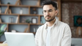 Young adult man wearing glasses working on laptop sitting on sofa in living room at home office, focused on task. Handsome male freelancer using computer application, texting a client, chatting online - Powered by Shutterstock - Get 15% off with code: PIKWIZARD15