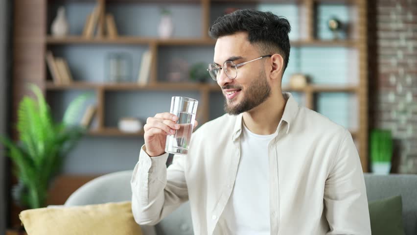 Young adult man drinks water from a glass in the living room at home. Happy smiling handsome male with feeling relieved, enjoying a clean cool drink shows thumb up looking at the camera. Close up