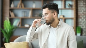 Young adult man drinks water from a glass in the living room at home. Happy smiling handsome male with feeling relieved, enjoying a clean cool drink shows thumb up looking at the camera. Close up - Powered by Shutterstock - Get 15% off with code: PIKWIZARD15