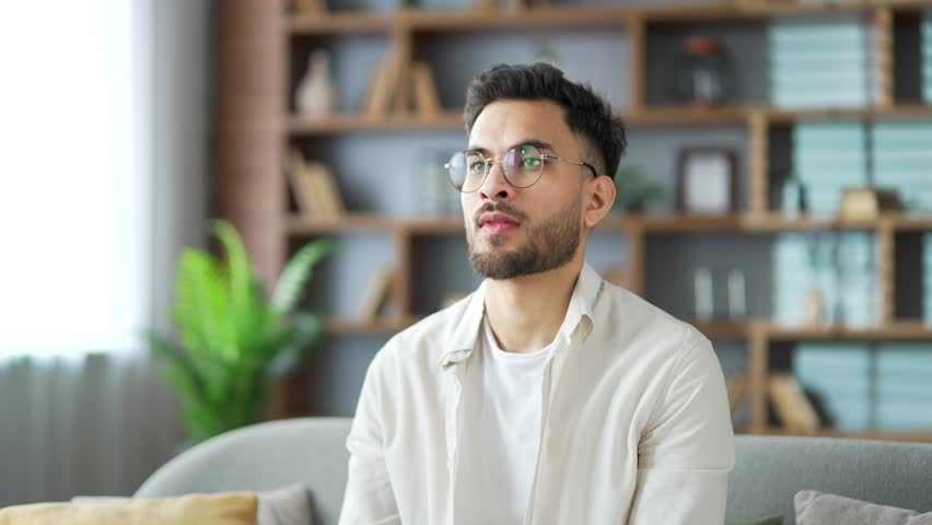 Smiling handsome bearded man relaxing with his eyes closed and his hands behind his head while sitting on sofa in living room at home. Positive happy male freelancer in shirt resting after work