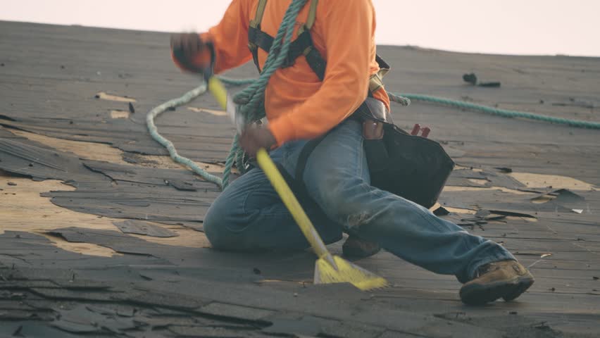Roofer using tools to strip worn-out shingles while secured with a safety harness.