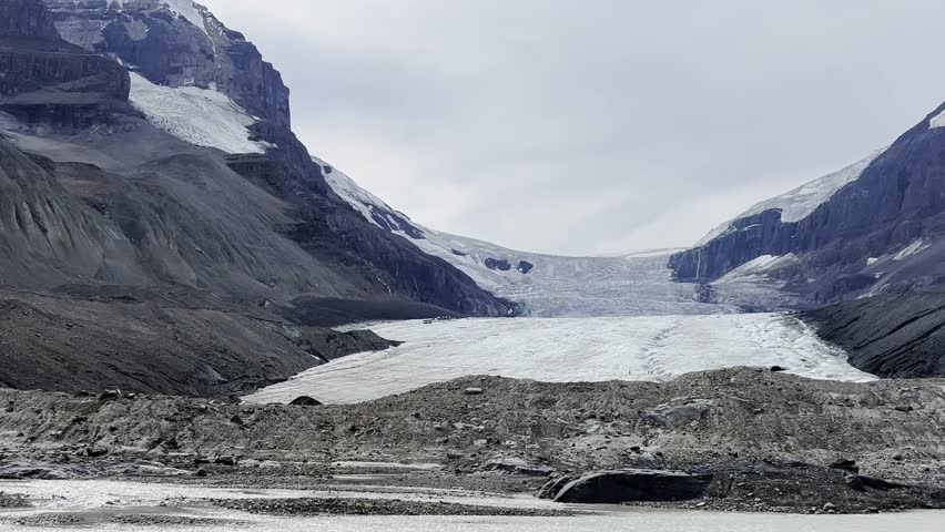 Striking view of Athabasca Glacier, part of Columbia Icefield in Jasper National Park, Alberta, Canada, with icy expanse flanked by rugged, rocky peaks under cloudy sky, Canada, zoom out