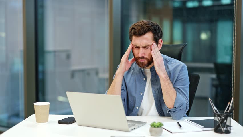 Stressed businessman with headache working on laptop in modern office. Tired man massages temples, feels unhealthy, exhausted. Overworked male having migraine, bad blurry vision.