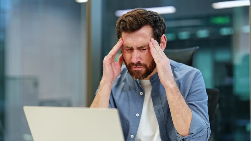 Frustrated businessman experiencing a bad headache in a modern office. He rubs temples and has painful expression while working on laptop.