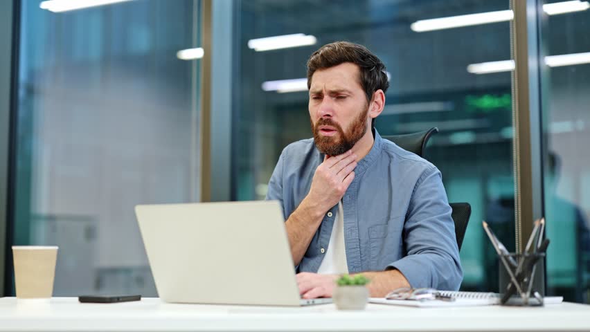 Unwell businessman with a sore throat sitting at workplace in modern office and working on laptop. Male employee in blue shirt having painful throat infection, tonsillitis or flu symptoms.