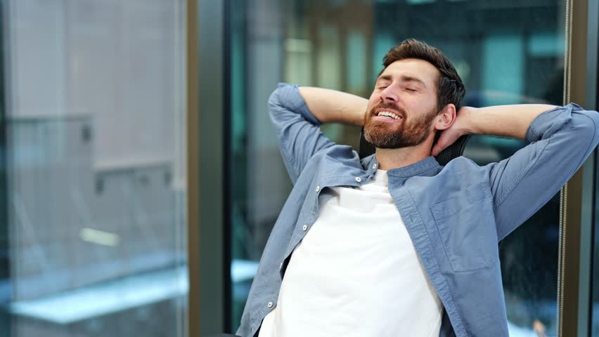 Successful businessman closes laptop, leans back, relaxes in office chair, puts hands behind head, and enjoys work done. Modern office interior.
