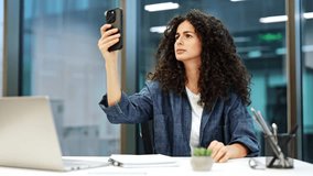 Focused businesswoman with curly hair having video call in modern office. Serious woman using smartphone for online meeting at workplace. - Powered by Shutterstock - Get 15% off with code: PIKWIZARD15