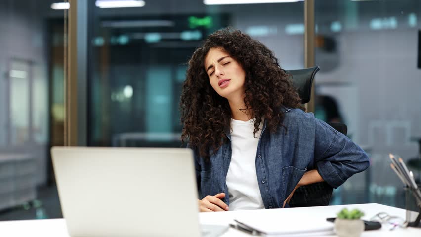 Curly haired businesswoman working on a laptop feels sudden lower back pain. Exhausted employee sitting in the office chair suffers from backache. Health problems at work concept.