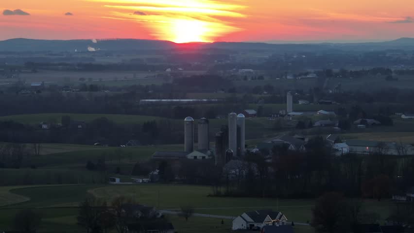 Farmstead with silo storage, barn and stables in american rural area. Sunset time behind hills during foggy and misty evening in USA. Aerial zoom wide shot. Establishing scene.