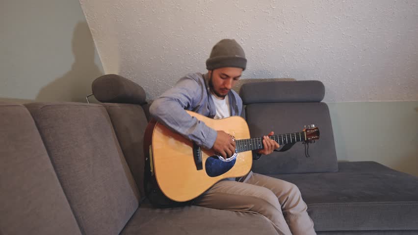 Musician sits indoors on a cozy couch, strumming an acoustic guitar in a warmly lit living room. The video captures their relaxed posture and the soothing tones of the music