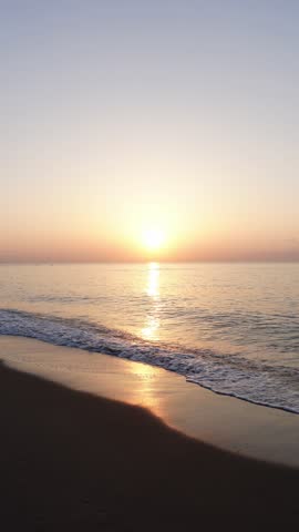 Beautiful sunrise or sunset from a sandy beach where the waves of a calm Mediterranean sea arrive. The sun rising over the horizon line that separates the water from a clear sky. Vertical video